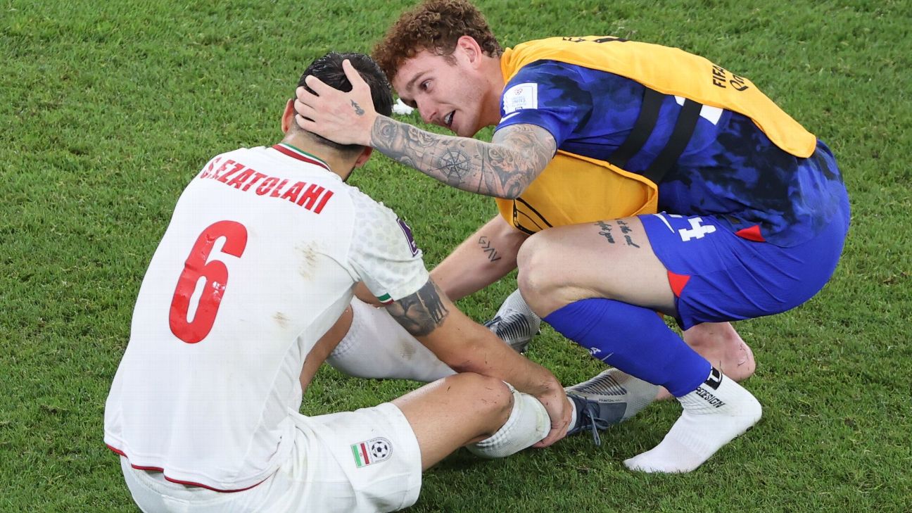 Saied Ezatolahi, in a white and red Iran uniform, sits on the grass dejected as American Josh Sargent crouches and puts his arm around him, consoling him