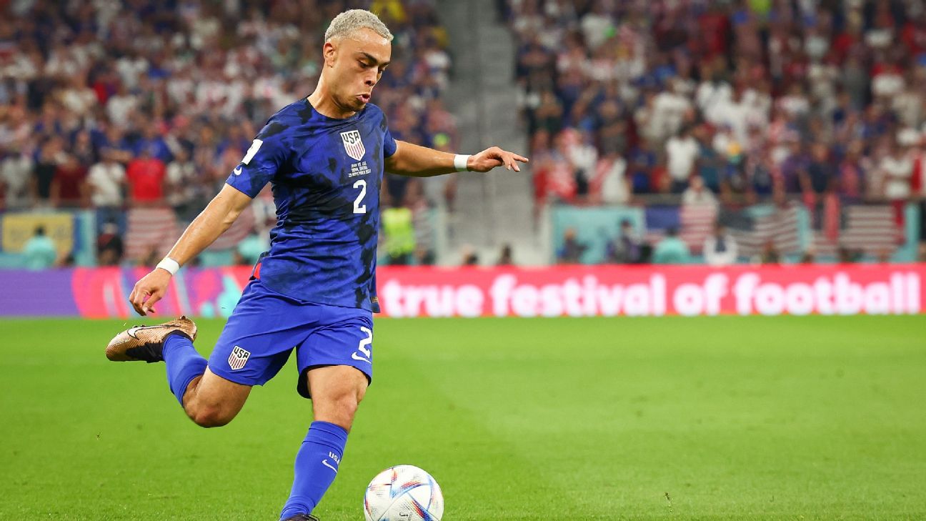 Sergino Dest wearing a blue USA uniform kicks the ball on a green grass soccer field