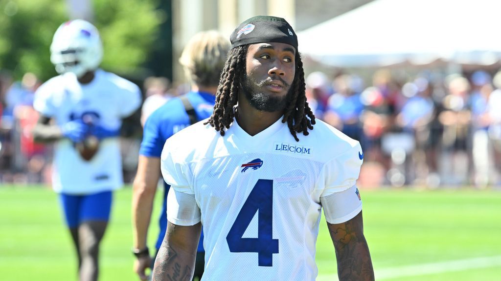 A football player wearing a white jersey with the number 4 and the Buffalo Bills logo stands on a grassy field. He has long dreadlocks and is wearing a black headband with the team's logo. In the background, another player in similar attire is slightly out of focus, and several people are gathered, suggesting a practice or training session.