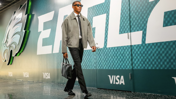 Philadelphia Eagles WR DeVonta Smith walks through the stadium corridor wearing a gray jacket, black trousers, sunglasses, and polished shoes while carrying a black leather bag.